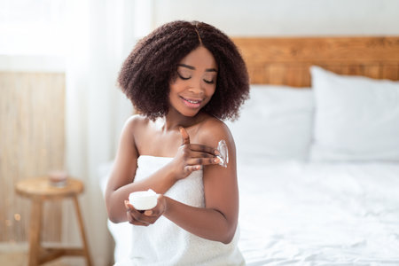 Lovely black lady wrapped in towel applying body cream on her shoulder after bath in bedroomの写真素材