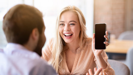Cheerful young woman showing her boyfriend smartphone with empty screen at coffee shop, mockup for designの写真素材