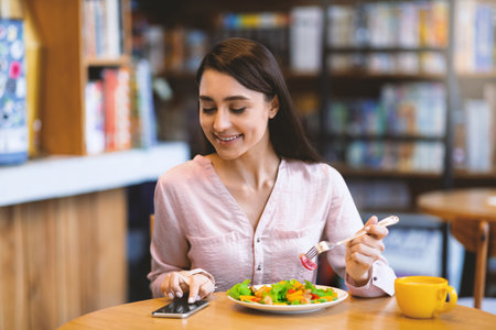 Happy young arab woman texting on smartphone while eating salad in cafe, using an application to send an smsの写真素材