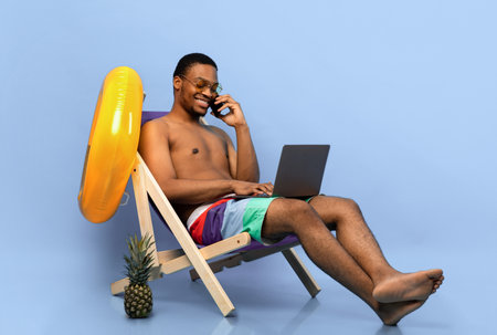 Online work on summer vacation. Black guy with laptop speaking on smartphone while sitting in lounge chairの写真素材