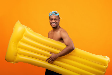 Smiling black man in snorkeling mask carrying inflatable lilo, going to beach on yellow studio backgroundの写真素材
