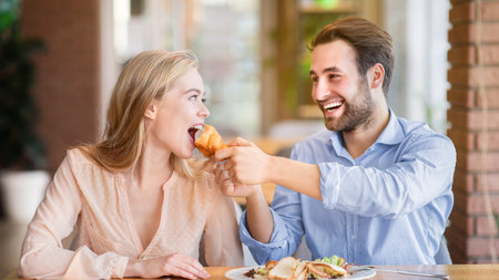 Affectionate young man feeding tasty croissant to his girlfriend at cozy cafeの写真素材