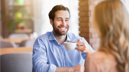 Smiling millennial guy drinking coffee with his girlfriend at cafe, having fun conversation during breakfastの写真素材