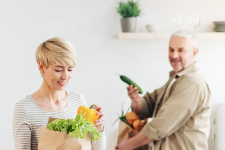Senior couple arriving from supermarket with grocery bags and unpacking vegetables in kitchen in morning for cookingの写真素材
