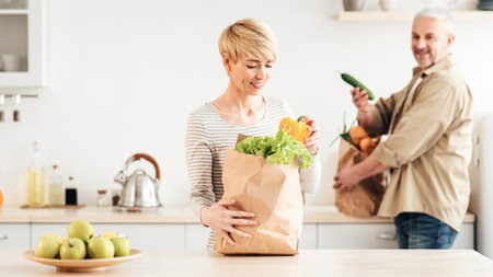 Senior couple arriving from supermarket with grocery bags and unpacking vegetables in kitchen in morning for cookingの写真素材