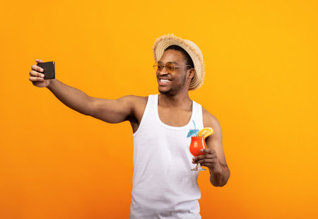 Im on vacation. Smiling black guy in summer wear taking selfie with tropical fruit cocktail on yellow studio backgroundの写真素材