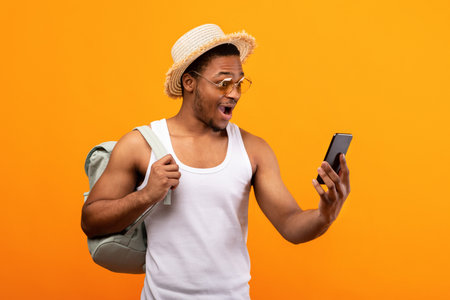 Black guy with backpack looking at smartphone in excitement, booking last minute travel tour online, yellow backgroundの写真素材
