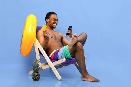 Smiling black man using credit card and smartphone for online shopping while chilling in lounge chair on blue backgroundの写真素材