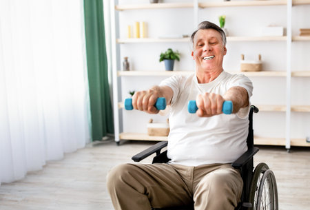 Joyful disabled man in wheelchair making rehabilitation exerises with dumbbells at retirement homeの写真素材