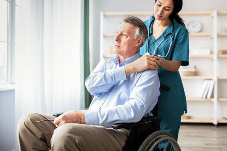 Young doctor holding elderly disabled males hand, giving him support at retirement home