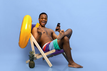 Excited black man holding credit card and smartphone, shopping online in lounge chair on blue studio backgroundの写真素材