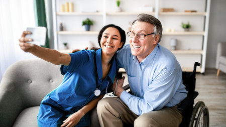 Cheerful impaired senior man in wheelchair taking selfie with young female doctor at retirement homeの写真素材