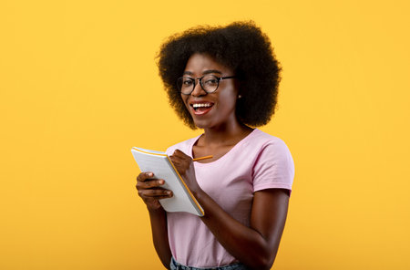 Checklist concept. Smart black female student wearing glasses, taking notes in copybook on yellow studio backgroundの写真素材