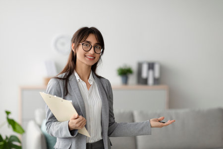 Happy middle eastern female psychologist with clipboard showing something with hand, smiling at camera, copy spaceの写真素材