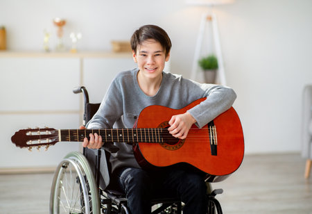 Happy disabled teen boy in wheelchair playing guitar, using musical instrument indoors. Teenage hobbies and pastimesの写真素材