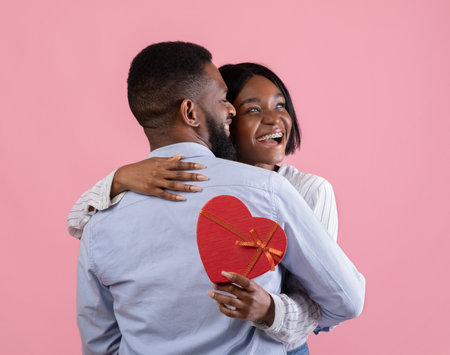 Excited black woman holding heart shaped gift box and hugging her boyfriend on pink studio backgroundの写真素材