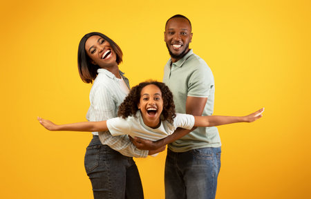 Family bonding concept. Joyful black man and woman holding daughter on hands, having fun over yellow studio wallの写真素材
