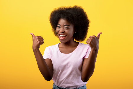 Best choice. Happy african american lady showing thumbs up gesture with both hands, posing on yellow studio wallの写真素材