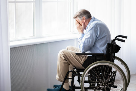 Sad and lonely senior disabled man in wheelchair crying near window at home. Depression in elderly impaired peopleの写真素材