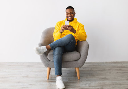 African Guy Using Smartphone Sitting In Chair Over Gray Wallの写真素材