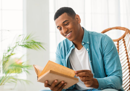 Smiling smart millennial african american guy reading book, sit on chair, enjoy comfort and free timeの写真素材