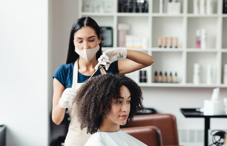 Hairdresser in protective mask cutting hair of curly african american client in beauty salonの写真素材