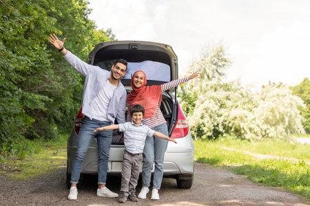 Glad cheerful emotional arab male, female in hijab and little boy waving their hands near car trunk, enjoy journeyの写真素材