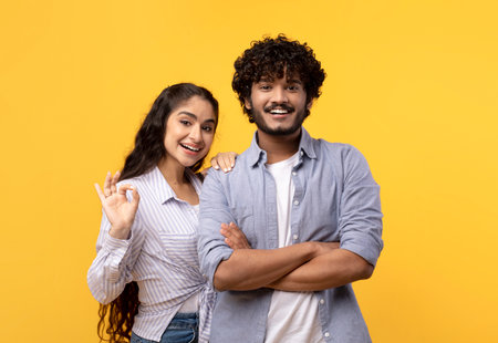 Portrait of happy indian couple embracing, woman showing okay gesture and smiling at camera over yellow backgroundの写真素材
