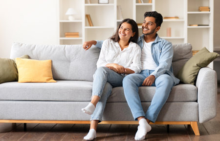 Happy dreamy indian couple looking aside at free space while sitting on sofaの写真素材