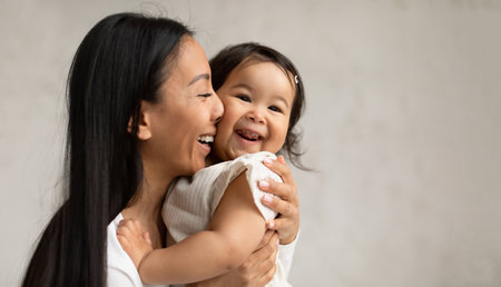 Joyful Asian Mom Hugging Baby Daughter Over Gray Wall Indoorの写真素材