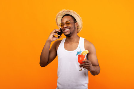 Positive black guy in trendy outfit holding tropical fruit cocktail and speaking on smartphone on yellow backgroundの写真素材
