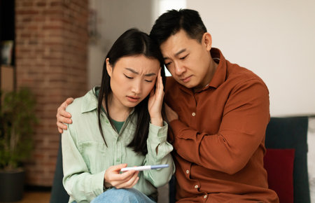 Upset mature asian husband comforting his depressed young wife with negative pregnancy test, sitting on sofa at homeの写真素材