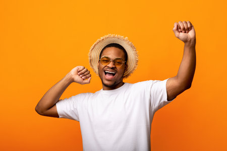 Handsome black man in sunglasses, t-shirt and straw hat raising his hands up, dancing on yellow studio backgroundの写真素材