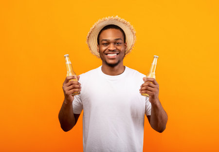 Cheerful young African American man holding two bottles of beer and smiling at camera on yellow studio backgroundの写真素材