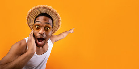 Summer sale. Shocked black guy in straw hat pointing at empty space on yelow studio backgroundの写真素材