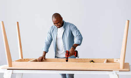 Carpentry service. Black handyman drilling wooden planks of table over light wall, free spaceの写真素材