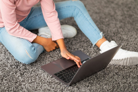 Browsing internet. Unrecognizable black girl sitting on floor carpet in living room, using laptop computer, cropの写真素材