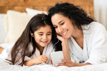 Smiling mom and little girl laying on bed, using smartphoneの写真素材