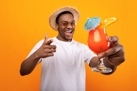 Carefree black guy in casual wear holding tropical cocktail and pointing at camera on yellow studio backgroundの写真素材
