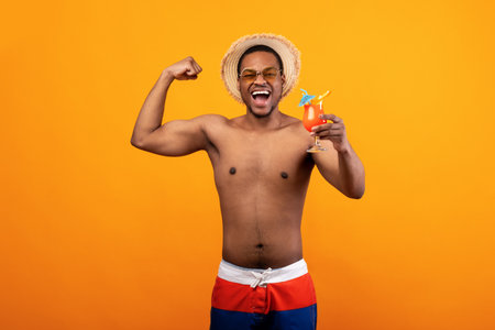 Emotional black man with naked chest drinking healthy tropical fruit cocktail, feeling strong, showing bicep musclesの写真素材
