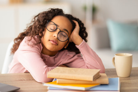 Tired teenager. African-american girl sleeping while studying with paper books, wearing glassesの写真素材