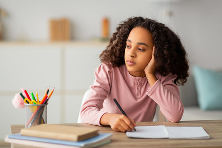 Thoughtful black teen girl writing in notepad, doing homework and looking away, sitting at table...