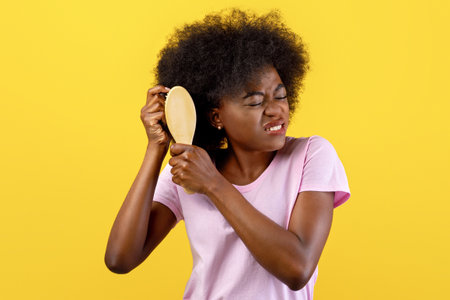 Annoyed african american woman having problem brushing her bushy curly hair on yellow studio backgroundの写真素材