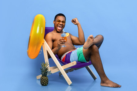 Full length of excited black man drinking tropical cocktail in lounge chair, gesturing YES over blue backgroundの写真素材