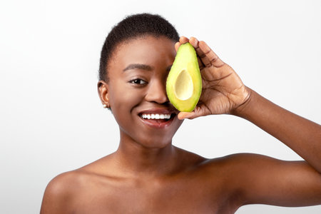 Happy african american young woman holding fresh avocado halves and covering eye, smiling to camera...