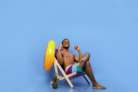 Relaxed black guy in swimwear chilling in lounge chair, pointing upwards at empty space over blue...