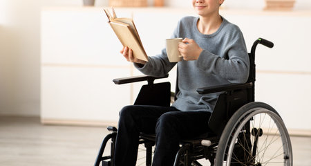 Positive impaired teenager in wheelchair drinking tea and reading interesting book at homeの写真素材