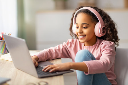 Happy teenage girl chatting on laptop, wearing wireless headphones, relaxing sitting at desk at home
