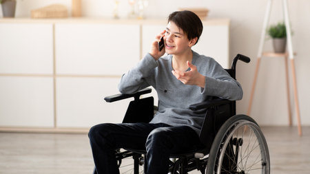 Cheerful teenager in wheelchair having phone conversation with friend at homeの写真素材