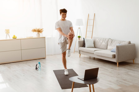 Asian guy doing standing knee to chest exercise using laptopの写真素材